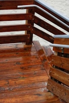 Wooden deck with a series of stairs and railings. The wood is richly colored and appears freshly varnished, with a glossy finish. The grain of the wood is clearly visible, and there are metal accents on the railings. The sandy ground is visible in the background.