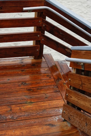 Wooden deck with a series of stairs and railings. The wood is richly colored and appears freshly varnished, with a glossy finish. The grain of the wood is clearly visible, and there are metal accents on the railings. The sandy ground is visible in the background.