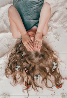 A person lies on a textured surface with hands covering their eyes. Long, light brown hair is spread out and decorated with flowers and butterflies. The overall scene has a gentle and whimsical aesthetic.