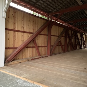 A wooden covered structure with a slanted roof, supported by thick wooden beams that form a series of diagonal and vertical patterns. The ground is covered with planks, and the lighting appears natural, coming from openings on the structures.
