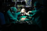 A team of surgeons in green surgical attire performs a procedure in an operating room. They focus intently on the task, with one of them carefully holding a newborn baby. The dim lighting creates a dramatic atmosphere, while surgical tools and equipment are visible on the table.