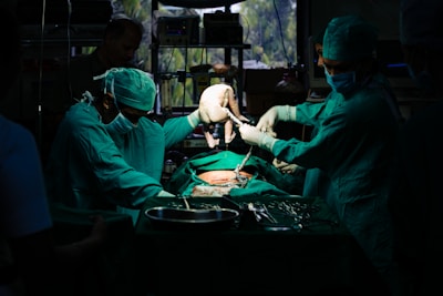 A team of surgeons in green surgical attire performs a procedure in an operating room. They focus intently on the task, with one of them carefully holding a newborn baby. The dim lighting creates a dramatic atmosphere, while surgical tools and equipment are visible on the table.