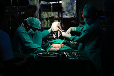 A team of surgeons in green surgical attire performs a procedure in an operating room. They focus intently on the task, with one of them carefully holding a newborn baby. The dim lighting creates a dramatic atmosphere, while surgical tools and equipment are visible on the table.