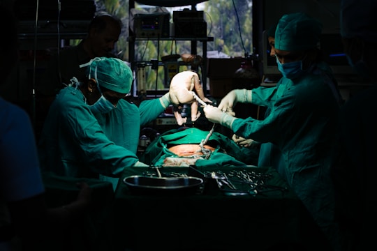 A team of surgeons in green surgical attire performs a procedure in an operating room. They focus intently on the task, with one of them carefully holding a newborn baby. The dim lighting creates a dramatic atmosphere, while surgical tools and equipment are visible on the table.