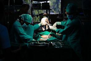 A team of surgeons in green surgical attire performs a procedure in an operating room. They focus intently on the task, with one of them carefully holding a newborn baby. The dim lighting creates a dramatic atmosphere, while surgical tools and equipment are visible on the table.