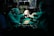 A team of surgeons in green surgical attire performs a procedure in an operating room. They focus intently on the task, with one of them carefully holding a newborn baby. The dim lighting creates a dramatic atmosphere, while surgical tools and equipment are visible on the table.