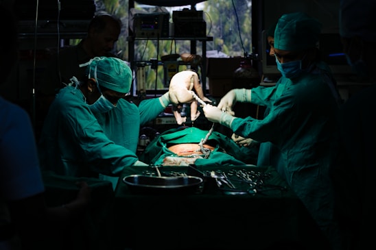 A team of surgeons in green surgical attire performs a procedure in an operating room. They focus intently on the task, with one of them carefully holding a newborn baby. The dim lighting creates a dramatic atmosphere, while surgical tools and equipment are visible on the table.
