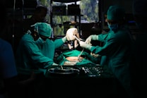A team of surgeons in green surgical attire performs a procedure in an operating room. They focus intently on the task, with one of them carefully holding a newborn baby. The dim lighting creates a dramatic atmosphere, while surgical tools and equipment are visible on the table.