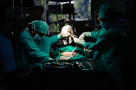 A team of surgeons in green surgical attire performs a procedure in an operating room. They focus intently on the task, with one of them carefully holding a newborn baby. The dim lighting creates a dramatic atmosphere, while surgical tools and equipment are visible on the table.