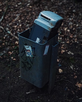 An old, rusted electronic device, possibly a cash register or electronic machine, is discarded inside a public trash bin. The bin is situated outdoors among dry leaves and twigs. Graffiti and stickers are visible on the bin’s surface, adding urban elements to the scene.