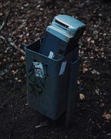 An old, rusted electronic device, possibly a cash register or electronic machine, is discarded inside a public trash bin. The bin is situated outdoors among dry leaves and twigs. Graffiti and stickers are visible on the bin’s surface, adding urban elements to the scene.