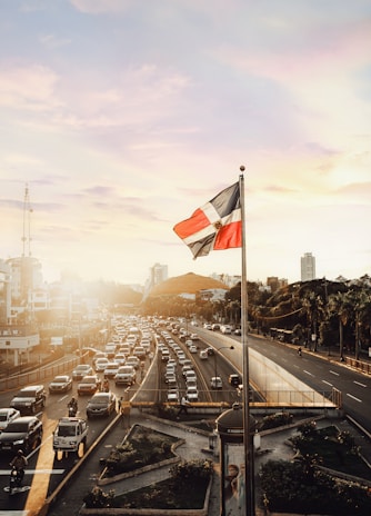 A bustling cityscape with a busy highway filled with cars during sunset. The Dominican Republic flag is prominently displayed on a pole in the foreground, gently swaying with the warm breeze. The sky is beautifully painted with hues of orange and pink as the sun sets in the background, casting a golden glow over the scene. Tall buildings and lush greenery line the roadsides, adding to the urban atmosphere.