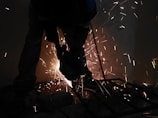Worker using a power sander on metal with sparks flying in a factory setting.