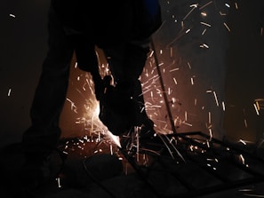 A worker uses a grinder on metal, creating bright sparks that illuminate the dimly lit environment.