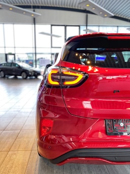 A bright red car is parked indoors in an expansive, well-lit showroom. The focus is on the rear, showcasing sleek taillights and a modern, aerodynamic design. Large windows allow natural light to flood the space, enhancing the polished look of the car. Other vehicles are visible in the background.