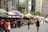 A bustling street market with vibrant, multicolored umbrellas covering various stalls. Shoppers are browsing through the items on display, including clothing, bags, and other merchandise. Buildings with signage in different languages line the street, adding to the lively atmosphere.