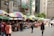 A bustling street market with vibrant, multicolored umbrellas covering various stalls. Shoppers are browsing through the items on display, including clothing, bags, and other merchandise. Buildings with signage in different languages line the street, adding to the lively atmosphere.