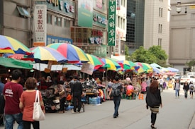 A bustling street market with vibrant, multicolored umbrellas covering various stalls. Shoppers are browsing through the items on display, including clothing, bags, and other merchandise. Buildings with signage in different languages line the street, adding to the lively atmosphere.