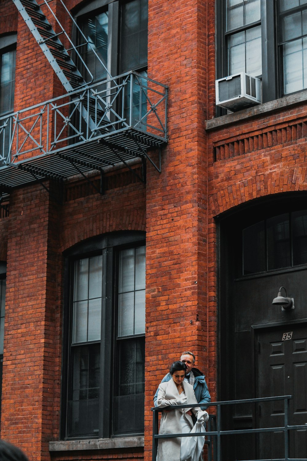 man in black jacket and blue denim jeans standing beside brown brick building during daytime