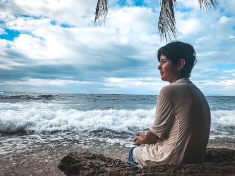 A person sits contemplatively on a rocky shore, gazing at the ocean waves. The sky is filled with clouds and a few palm leaves hang down, adding a tropical feel to the scene.