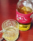 A bottle of Japanese plum-flavored whisky labeled 'Akashi Ume' with a red label sits on a wooden surface. Next to it is a glass mug filled with a similar colored liquid, likely the same whisky. The mug has an intricate design etched into the glass.
