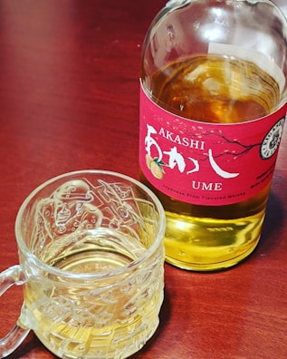 A bottle of Japanese plum-flavored whisky labeled 'Akashi Ume' with a red label sits on a wooden surface. Next to it is a glass mug filled with a similar colored liquid, likely the same whisky. The mug has an intricate design etched into the glass.