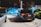 Multiple cars are parked in a dimly lit outdoor area, with one person standing nearby looking at a phone. The cars include a bright blue vehicle with its doors open and a black car with distinct red rear lights.
