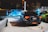 Multiple cars are parked in a dimly lit outdoor area, with one person standing nearby looking at a phone. The cars include a bright blue vehicle with its doors open and a black car with distinct red rear lights.