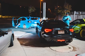 Multiple cars are parked in a dimly lit outdoor area, with one person standing nearby looking at a phone. The cars include a bright blue vehicle with its doors open and a black car with distinct red rear lights.