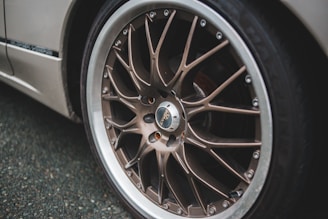 A close-up view of a car wheel featuring a detailed and stylish alloy rim design, with silver and bronze tones. The wheel appears to be on a parked vehicle with a dark textured road surface in the background.
