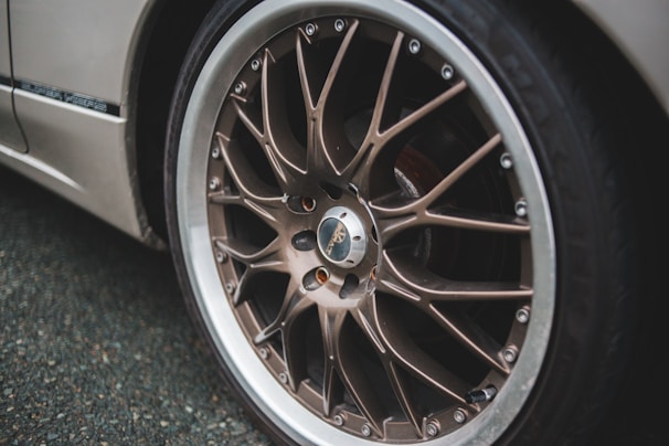 A close-up view of a car wheel featuring a detailed and stylish alloy rim design, with silver and bronze tones. The wheel appears to be on a parked vehicle with a dark textured road surface in the background.
