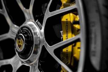 A close-up of a car wheel featuring a shiny metallic rim with a visible Porsche logo. Behind the spokes, a vibrant yellow brake caliper is prominently displayed. The image has a modern and sleek appearance, emphasizing luxury and performance.