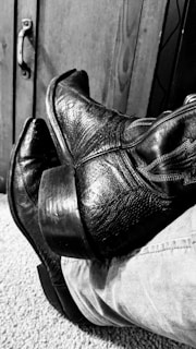 A pair of worn, textured leather cowboy boots rest on carpeted flooring beside a wooden cabinet with a metal handle. The image is in black and white, emphasizing the rugged and vintage quality of the boots.