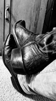 A pair of worn, textured leather cowboy boots rest on carpeted flooring beside a wooden cabinet with a metal handle. The image is in black and white, emphasizing the rugged and vintage quality of the boots.