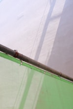 A metal pipe extends diagonally across the image, casting shadows on a light-colored wall. Below, a semi-transparent green sheet, possibly a tarp or covering, is draped and secured with metal grommets.