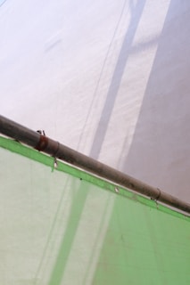 A metal pipe extends diagonally across the image, casting shadows on a light-colored wall. Below, a semi-transparent green sheet, possibly a tarp or covering, is draped and secured with metal grommets.