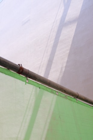 A metal pipe extends diagonally across the image, casting shadows on a light-colored wall. Below, a semi-transparent green sheet, possibly a tarp or covering, is draped and secured with metal grommets.