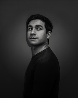 A black and white portrait of a person with short, dark hair looking slightly upwards towards the right. The background is dark, and the lighting highlights the facial features, creating a dramatic effect.