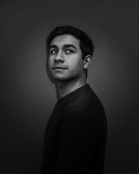 A black and white portrait of a person with short, dark hair looking slightly upwards towards the right. The background is dark, and the lighting highlights the facial features, creating a dramatic effect.