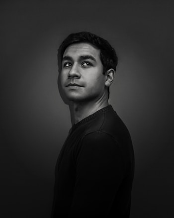 A black and white portrait of a person with short, dark hair looking slightly upwards towards the right. The background is dark, and the lighting highlights the facial features, creating a dramatic effect.