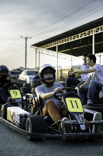 A person wearing a helmet is seated in a go-kart with number 11. Another go-kart numbered 9 is visible nearby. Three people are sitting on a bench in the background beneath a metal shelter. The setting is outdoors near dusk with a clear sky and some electrical poles in view.