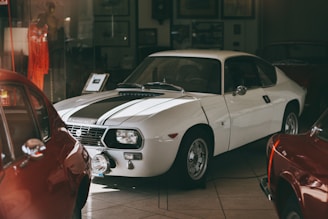A classic white sports car is prominently displayed in a dimly lit showroom. The car's sleek design features a black racing stripe running along the hood. Surrounding the main vehicle are other classic cars, partially visible, with their colorful bodies reflecting light. The scene includes various framed pictures and documents on the walls, hinting at a vintage automotive theme.