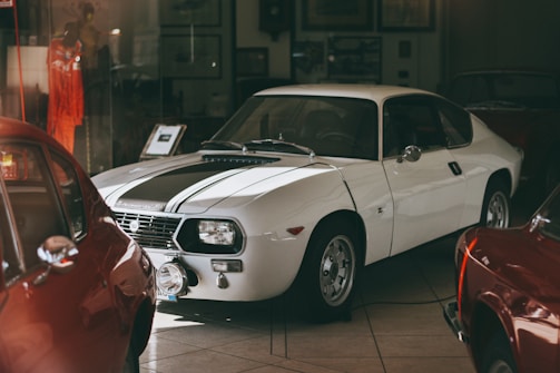 A classic white sports car is prominently displayed in a dimly lit showroom. The car's sleek design features a black racing stripe running along the hood. Surrounding the main vehicle are other classic cars, partially visible, with their colorful bodies reflecting light. The scene includes various framed pictures and documents on the walls, hinting at a vintage automotive theme.