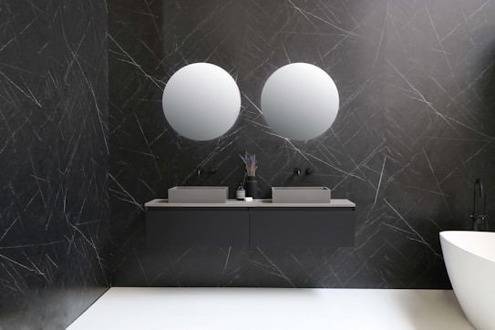 A modern bathroom features two round mirrors mounted on a dark textured wall with a marble pattern. Below the mirrors, there are two rectangular sinks with a minimalist design, accompanied by matching dark faucet fixtures. A vase with decorative items and a small container are placed on the countertop. To the right, a sleek white bathtub adds contrast to the ambiance.