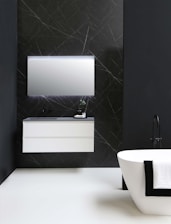 A modern bathroom features a sleek, white floating vanity with a mirror above, set against a dark wall with subtle line patterns. A standalone bathtub with a black faucet is adorned with a draped black and white towel.