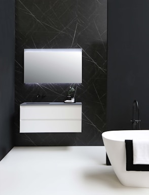 A modern bathroom features a sleek, white floating vanity with a mirror above, set against a dark wall with subtle line patterns. A standalone bathtub with a black faucet is adorned with a draped black and white towel.