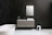 A modern bathroom with a sleek, floating vanity featuring a rectangular mirror above. The white countertop has a small plant and a soap dish. The bathtub is freestanding with a minimalist design, next to a stack of folded towels in a basket. The wall is dark with subtle lines, adding texture to the space.