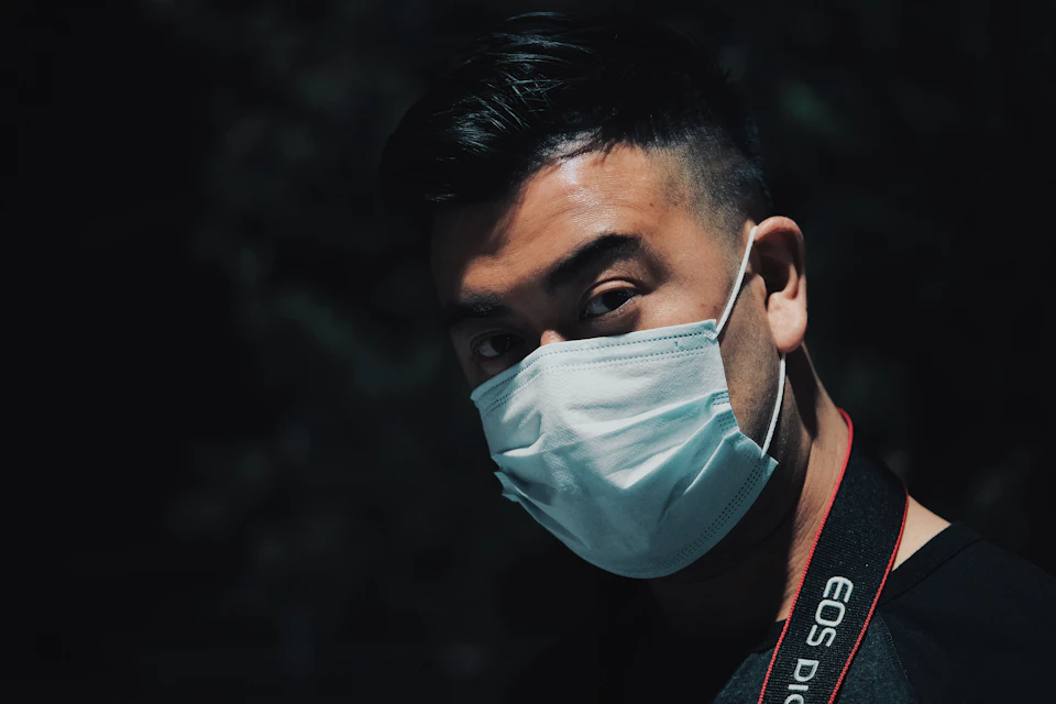 The image shows a man wearing a white surgical mask with red straps, standing against a dark background. The man's face is partially obscured by the mask, and he appears to be looking directly at the camera.