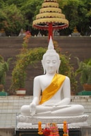 A large white Buddha statue is seated in a traditional meditation pose with a serene expression, draped in a golden sash. An ornate multi-tiered umbrella stands above the statue, and the surroundings include potted plants and greenery.