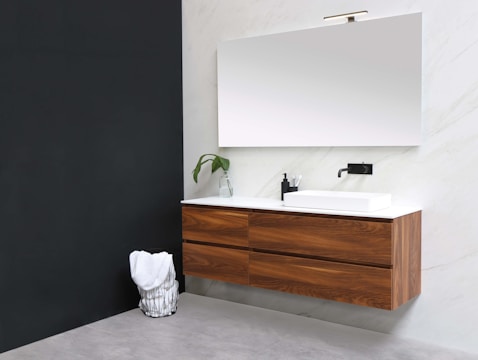 A modern bathroom with a minimalistic design featuring a floating wooden vanity with sleek drawers. A large frameless mirror is mounted above the vanity, reflecting light. Beside the sink is a black faucet and a small black container holding toothbrushes. A potted plant in a glass vase adds a touch of greenery. The walls are a combination of dark and light colors, creating a contemporary look, and a wire basket holds a white towel on the floor.